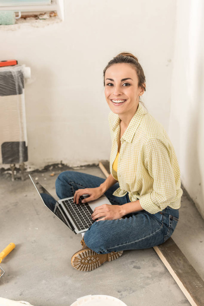 attractive young woman using laptop and smiling at camera during home improvement - Photo, Image