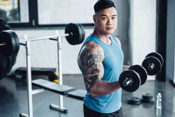 Konzentrierter junger asiatischer Sportler trainiert mit Kurzhanteln im Fitnessstudio und blickt in die Kamera - Foto, Bild