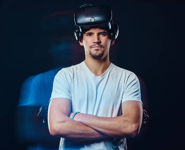 Jogador profissional com cerda vestida com camisa branca vestindo óculos de realidade virtual e mantém joysticks, posando com braços cruzados
. - Foto, Imagem