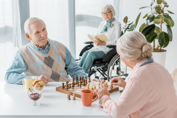 senior couple playing chess while senior woman sitting in wheelchair and reading book - Foto, afbeelding