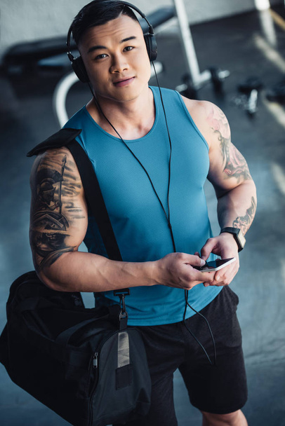 young asian sportsman using smartphone and listening to music at gym with sportive bag - Foto, imagen