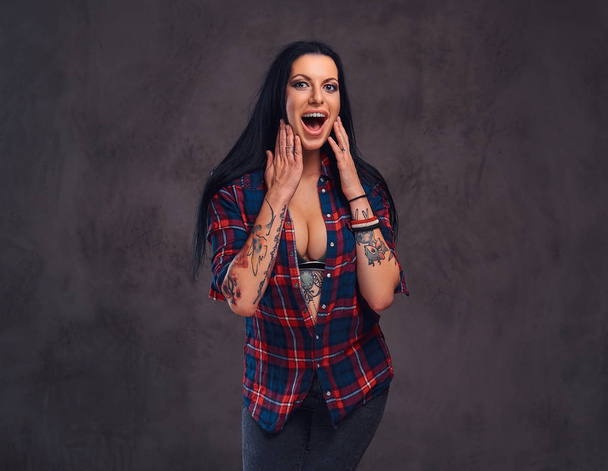 Surprised tattooed girl wearing a red unbuttoned checked shirt in a studio. - Photo, Image