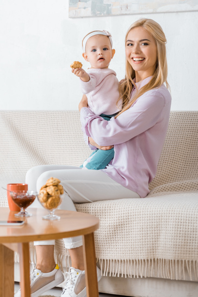 young woman sitting on sofa with small daughter holding cookie - Foto, Imagem