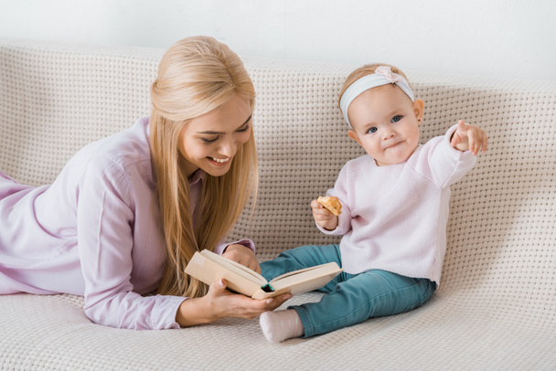 joven feliz madre acostado en sofá y lectura libro a pequeña hija mientras niño sosteniendo galleta
 - Foto, imagen
