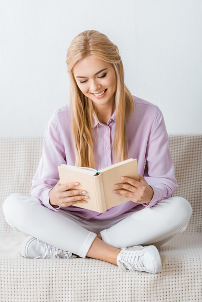 beautiful smiling woman reading book and sitting on sofa - Zdjęcie, obraz