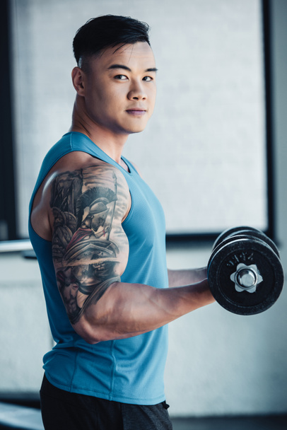 joven tatuado asiático deportista ejercitando con dumbbell en gimnasio y mirando cámara
 - Foto, Imagen