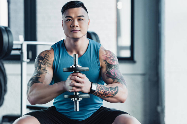 concentrated young asian sportsman exercising with dumbbell in gym and looking at camera - Photo, Image