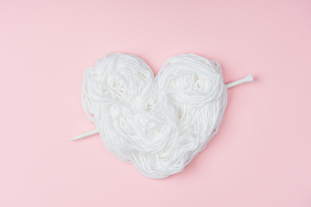 top view of yarn arranged in heart symbol and knitting needle on pink backdrop - Фото, изображение