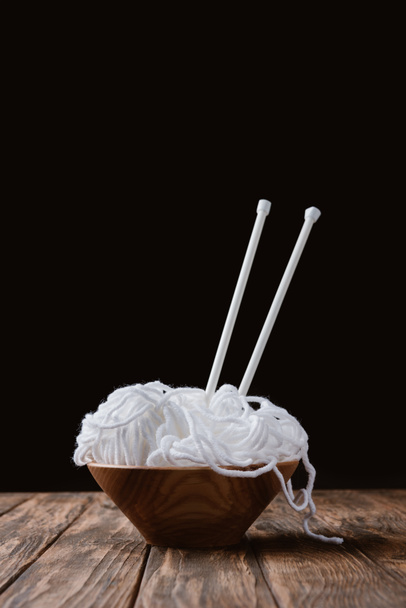 close up view of white yarn in bowl and knitting needles on wooden surface on black backdrop - Photo, Image