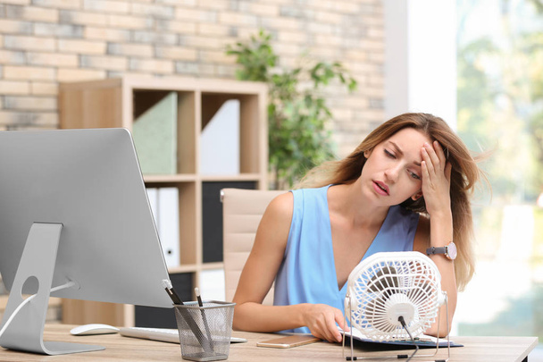 Businesswoman suffering from heat in front of small fan at workplace - 写真・画像