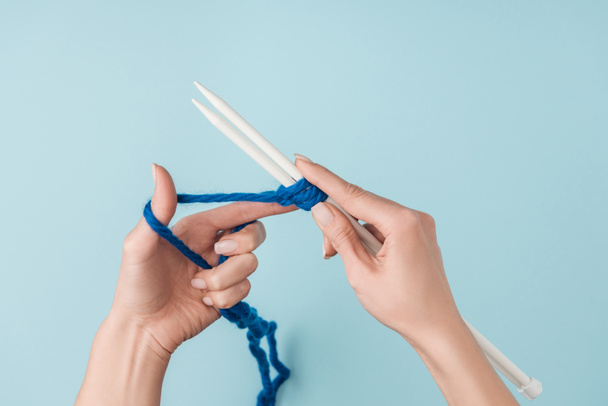 partial view of woman with blue yarn and white knitting needles knitting on blue backdrop - Fotografie, Obrázek