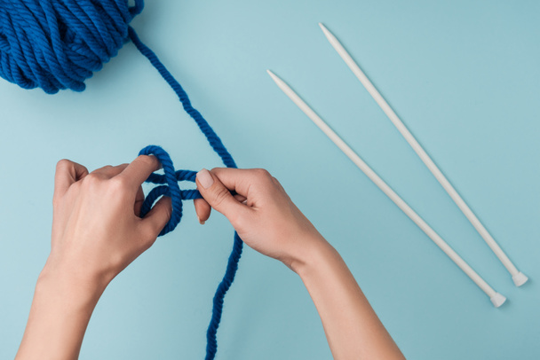 partial view of woman with blue yarn and white knitting needles knitting on blue backdrop - Фото, изображение