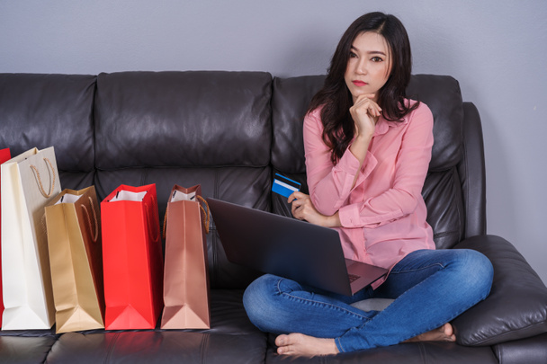 Frau denkt und nutzt Laptop für Online-Einkäufe mit Kreditkarte im Wohnzimmer - Foto, Bild