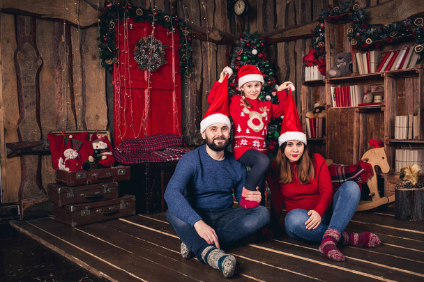 Щаслива сім'я в різдвяних капелюхах сидить у кімнаті в сільському стилі. Дочка виховує шапки батькам
. - Фото, зображення