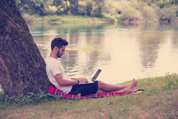 Ένας νεαρός ελεύθερος επαγγελματίας χρησιμοποιώντας έναν φορητό υπολογιστή ενώ λειτουργεί στην πανέμορφη φύση κάτω από το δέντρο στην όχθη του ποταμού - Φωτογραφία, εικόνα