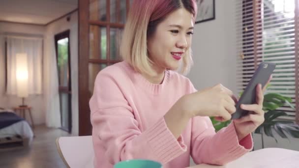 Young Asian woman using smartphone while lying on the desk in her living room. Happy female use phone for texting, reading, messaging and buying online at home. Lifestyle woman at home concept. - Imágenes, Vídeo