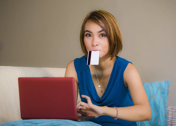 lifestyle portrait of young beautiful and happy girl  at home living room holding credit card using laptop computer for banking and online shopping smiling relaxed in e-business and internet commerce  - 写真・画像