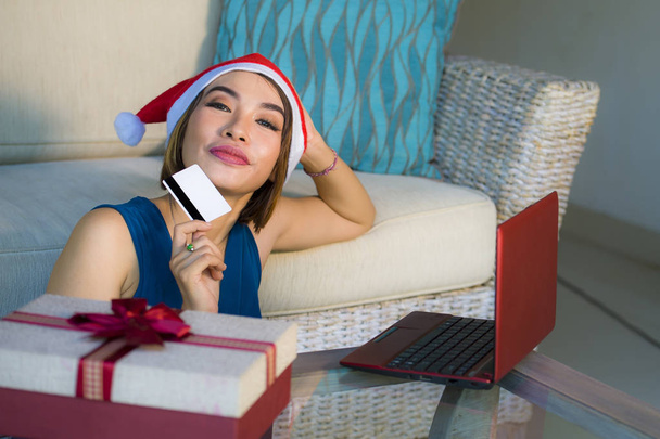 幸せで、美しい少女がクリスマス プレゼントのオンライン ショッピング、インターネット商取引に笑みを浮かべてクレジット カードで支払いのラップトップ コンピューターを使用してサンタの帽子でソファでリラックス自宅 - 写真・画像