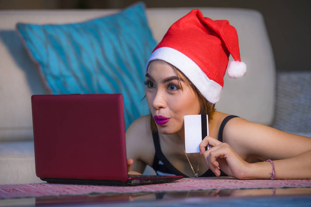 young happy and beautiful girl relaxed at home couch in Santa hat using laptop computer paying for Christmas present with credit card smiling in online shopping and internet commerce - 写真・画像