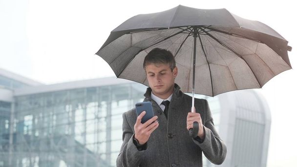 Πορτραίτο του όμορφος επιχείρηση άνθρωπος χρησιμοποιώντας το smartphone. Νεαρός επιχειρηματίας κρατώντας ομπρέλα και χρησιμοποιώντας ένα έξυπνο τηλέφωνο σε μια βροχερή ημέρα. - Φωτογραφία, εικόνα