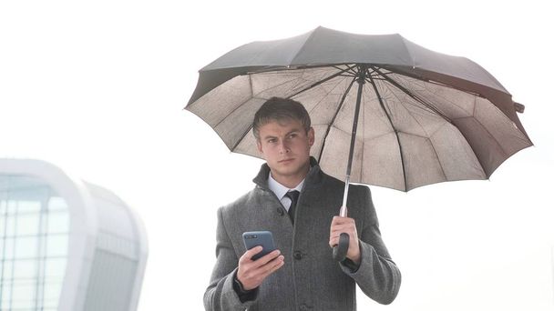 Portrait of handsome business man using smartphone. Young businessman holding umbrella and using a smart phone in a rainy day. - 写真・画像