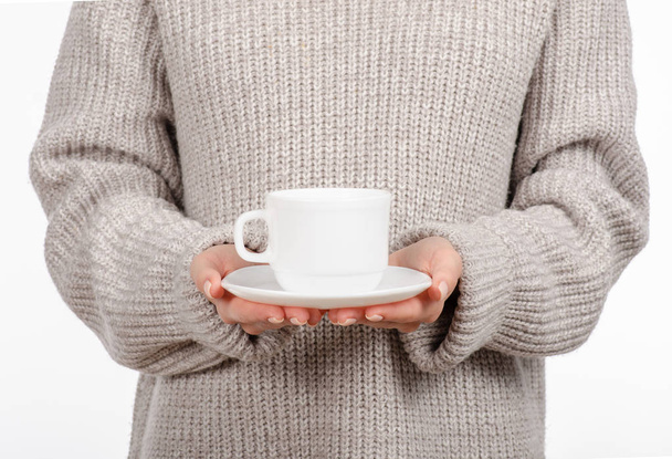 Young woman in sweater holding a white cup and saucer in hands, drinking hot coffee. - Fotó, kép