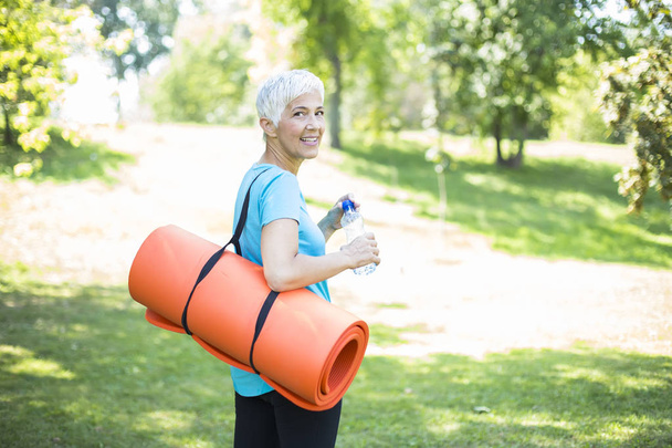 Retrato de mulher sênior mantém tapete de fitness em suas costas no parque e se preparando para o exercício
 - Foto, Imagem