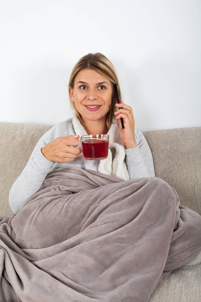Cheerful young woman relaxing on the sofa, drinking hot tea and talking on the phone with friend - Foto, Imagen