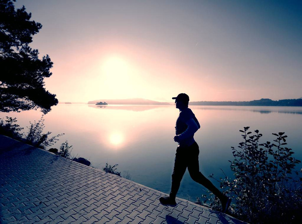 Alto hombre delgado está corriendo a lo largo del lago en el parque en el amanecer soleado. Niveles de agua brumosa
. - Foto, Imagen
