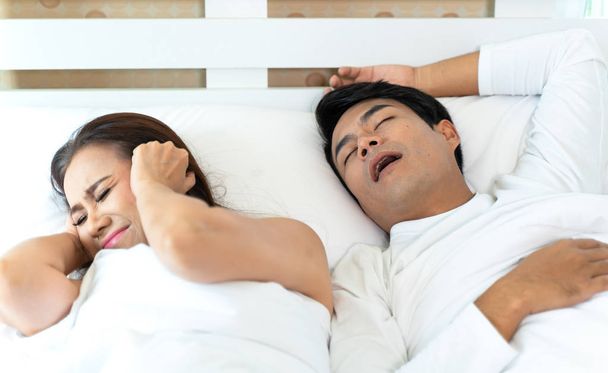Frau kann nicht schlafen und verdeckt ihre Ohren vor Lärm, während Mann zu Hause im Bett schnarcht - Foto, Bild