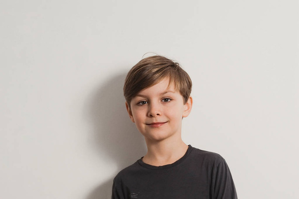 a closeup portrait of a smiling cute boy in a grey shirt, leaning on the white wall - Photo, Image
