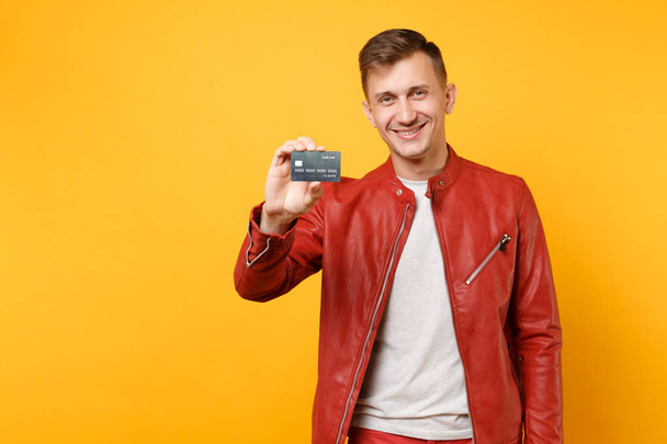 Portret vogue uśmiechający się przystojny młody mężczyzna w czerwonej skórzanej kurtce t-shirt przytrzymaj kartę kredytową banku na białym tle na najpopularniejsze żółte tło. Koncepcja życia szczere emocje ludzi. Powierzchnia reklamowa - Zdjęcie, obraz