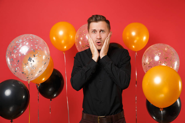 Bewildered young man in black classic shirt keeping hands on cheeks on bright red background air balloons. Valentine's International Women's Day, Happy New Year, birthday mockup holiday party concept - Foto, Imagem