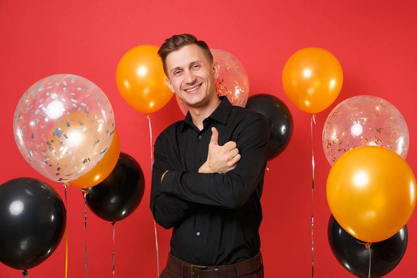 Young man in black classic shirt celebrating, holding hands folded, showing thumb up on bright red background air balloons. St. Valentine's Day, Happy New Year, birthday mockup holiday party concept - Photo, image