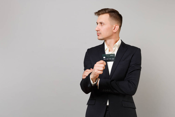 Portrait of young business man in classic black suit shirt looking aside holding credit bank card isolated on grey background in studio. Achievement career wealth business concept. Mock up copy space - Foto, immagini