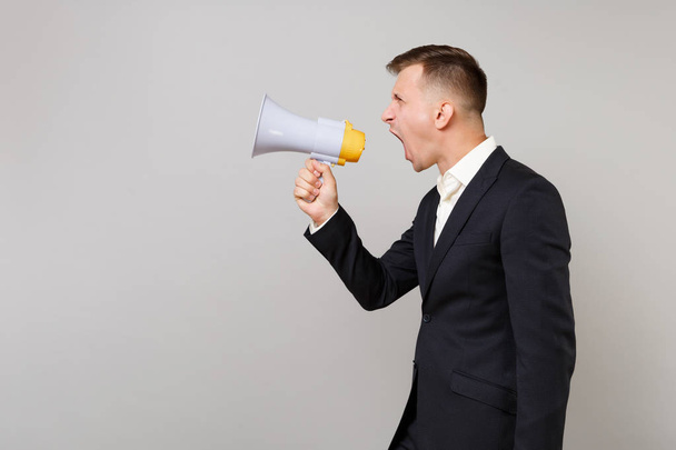 Side view of young business man in classic black suit and white shirt screaming on megaphone isolated on grey wall background in studio. Achievement career wealth business concept. Mock up copy space - Fotó, kép