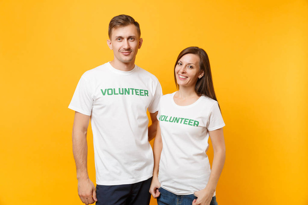 黄色の背景に分離されて書かれた碑文緑タイトル ボランティアの白い t シャツの若い同僚カップルの肖像。無料ボランティア、アシスタンスのヘルプ、チャリティー恵みチームワークの概念 - 写真・画像