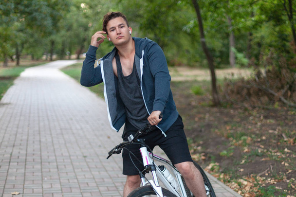 Attractive young caucasian man with dark hair bicycling in the park. Outdoors, autumn / fall park. Diversity people. Melancholy mood. Headphones (earphones, airpods), silver ring, white and rose bicycle.  - Photo, Image