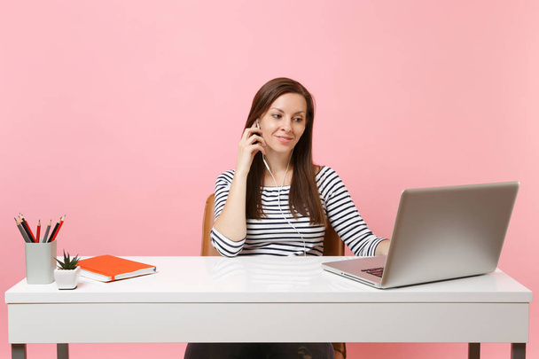 Mladými, krásnými žena s sluchátka poslouchat hudbu video volání sedět práci v kanceláři s moderní pc laptop izolované na pastelově růžové pozadí. Úspěch obchodní kariéru koncept. Kopírovat prostor - Fotografie, Obrázek