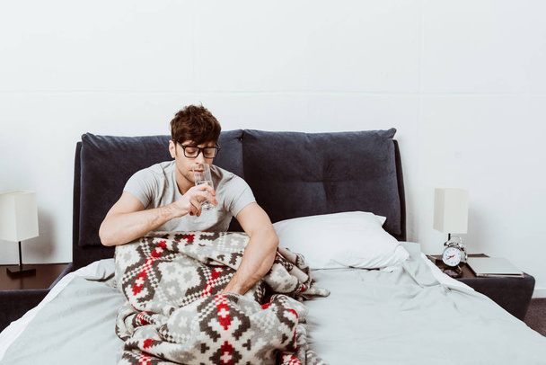 handsome young man in eyeglasses drinking water in bed at home - Фото, изображение