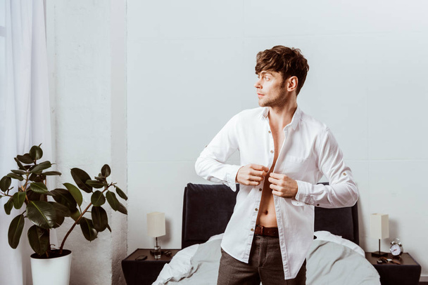 vista lateral de hombre de negocios guapo abotonando camisa blanca en el dormitorio en casa
 - Foto, Imagen