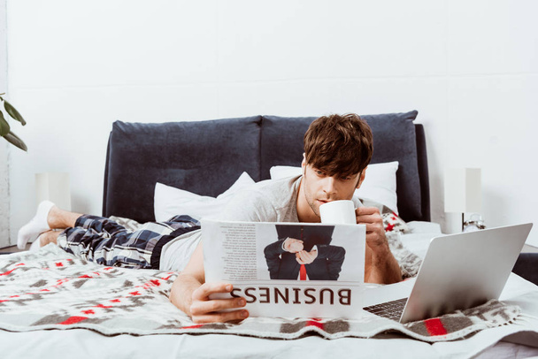 young man drinking coffee and reading business newspaper on bed with laptop at home  - Foto, immagini