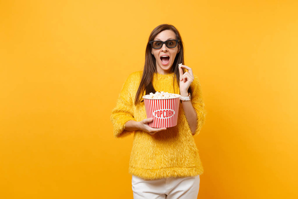 Surprised young girl in 3d imax glasses watching movie film and holding bucket of popcorn isolated on bright yellow background. People sincere emotions in cinema, lifestyle concept. Advertising area - Photo, Image
