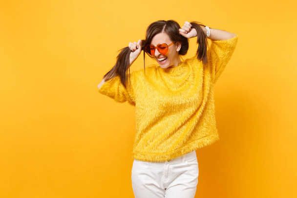 Laughing young woman in fur sweater, white pants, heart orange glasses holding hair like ponytails isolated on bright yellow background. People sincere emotions, lifestyle concept. Advertising area - Foto, Imagem