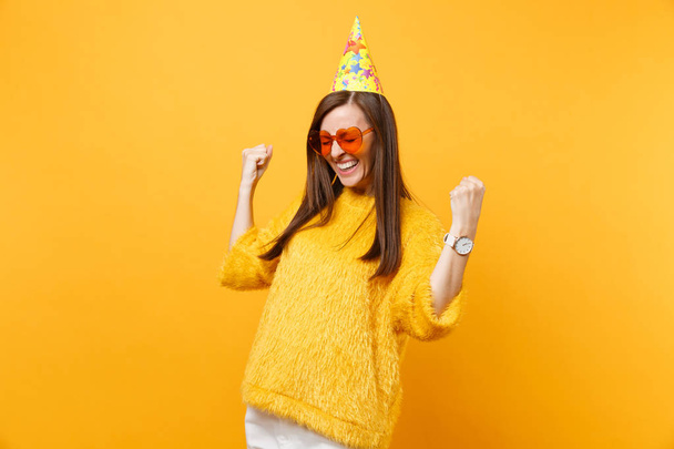 Радісна щаслива молода жінка в помаранчевих серцевих окулярах, день народження капелюх клейких кулаків, як переможець, насолоджується святом, святкуючи ізольовані на яскраво-жовтому тлі. Люди щирі емоції, спосіб життя
 - Фото, зображення