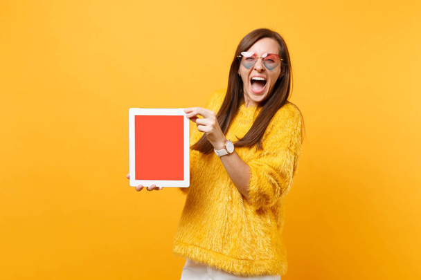 Overjoyed laughing young woman in heart glasses hold tablet pc computer with blank black empty screen isolated on bright yellow background. People sincere emotions lifestyle concept. Advertising area - Fotografie, Obrázek