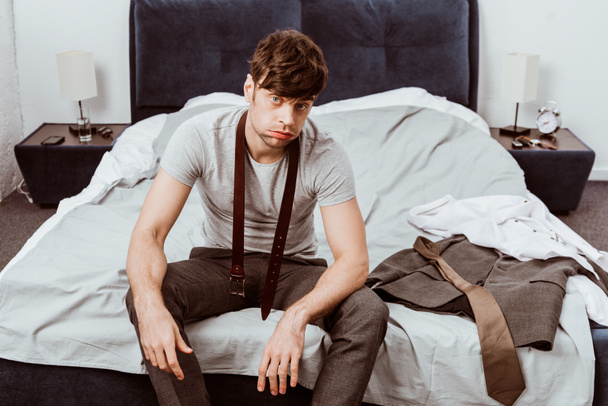 Müder junger Geschäftsmann mit Gürtel um den Hals blickt in die Kamera und sitzt zu Hause auf dem Bett - Foto, Bild