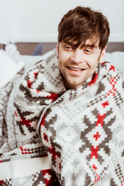 smiling sick young man wrapped in blanket sitting on bed at home - Foto, Imagem