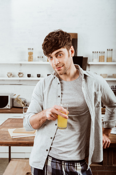 cheerful young man standing with orange juice in kitchen at home - Valokuva, kuva