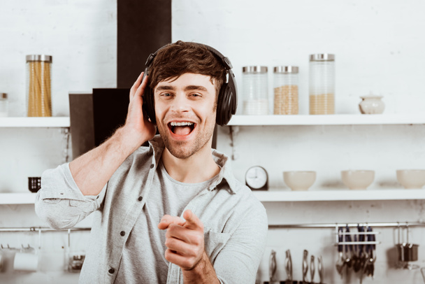 homem feliz em fones de ouvido piscando e apontando para a câmera na cozinha em casa
 - Foto, Imagem